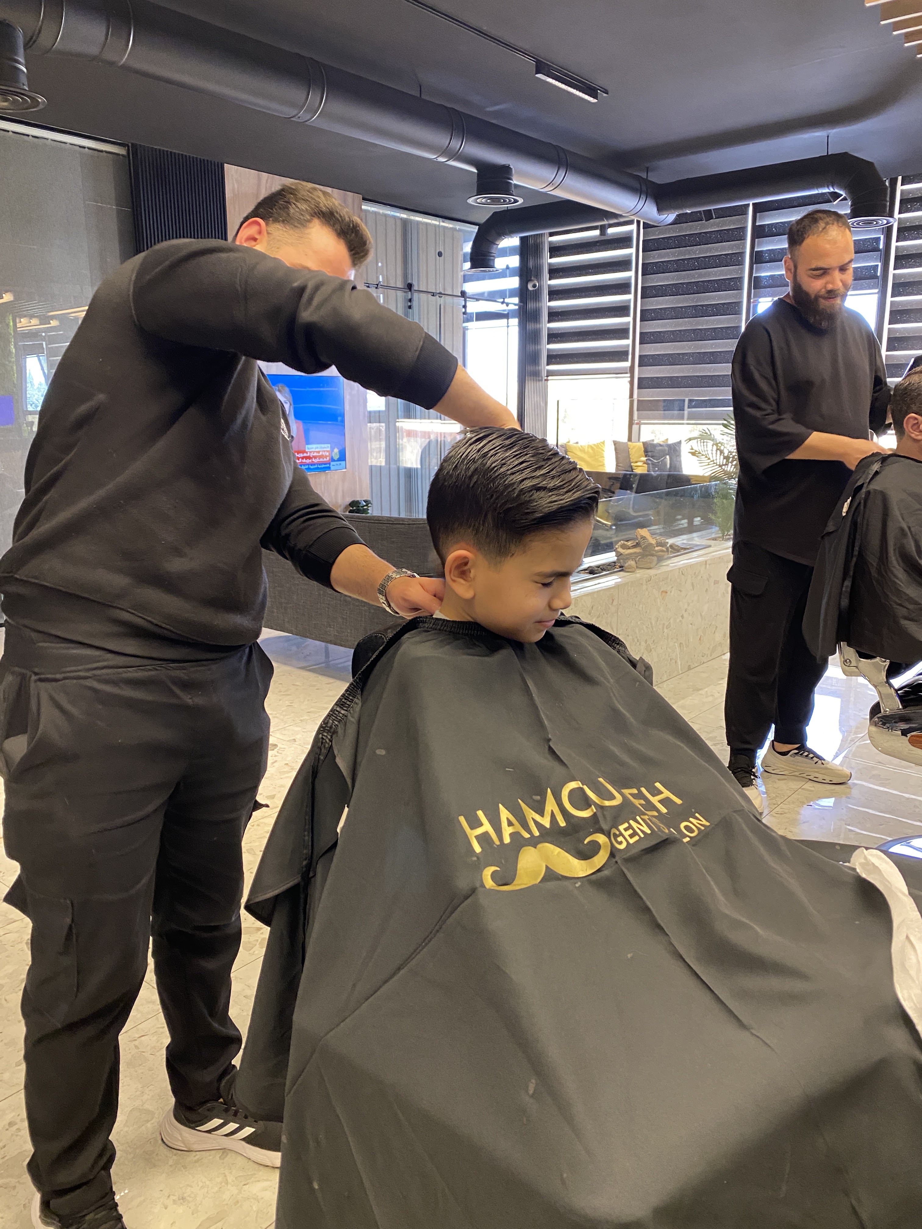 Barber finishing a child's haircut in a branded Hamoudeh Gent's Salon cape